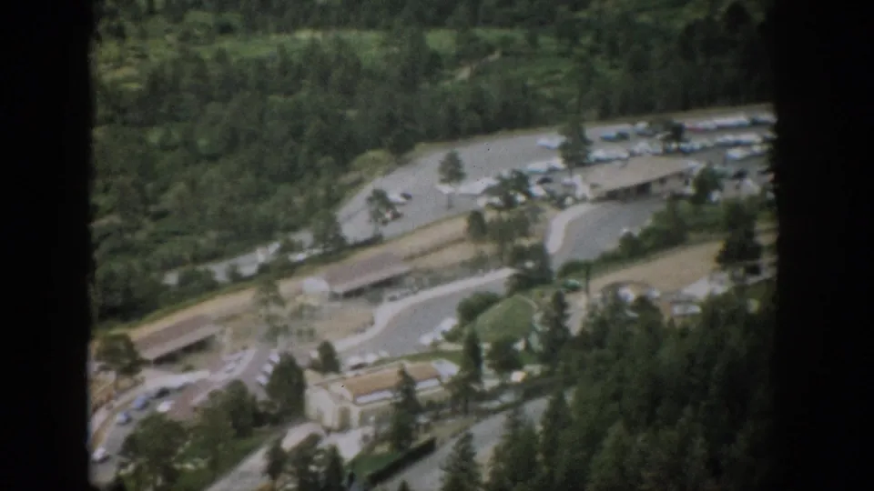 1959 Colorado home-movie aerial frame showing a road loop, parked cars, low town buildings, and dense pine forest in faded color.