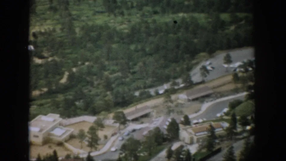 Soft 8mm aerial frame from Colorado showing forest, a curving road, small buildings, and cars near a mountain roadside area.
