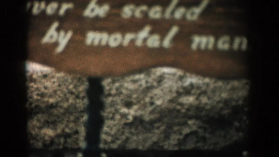 Close 8mm view of the wooden sign in Colorado, with white lettering reading in part “never be scaled by mortal men” over textured stone.