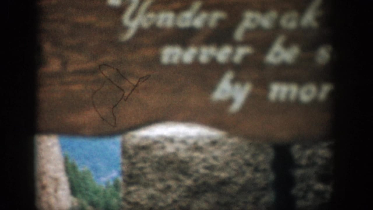 1959 Colorado home-movie frame with a rustic sign partly obscuring a mountain view and blue sky beyond.