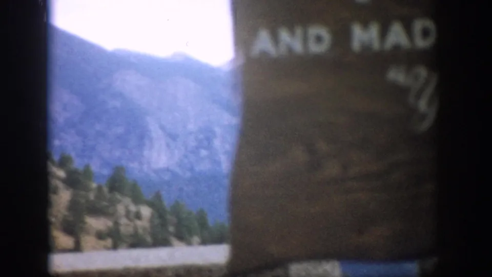 1959 Colorado 8mm frame shifting from the sign toward a hazy mountain slope with pine trees in the foreground.