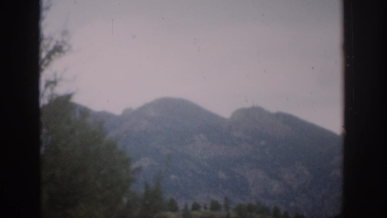 1959 Colorado mountain landscape filmed through a dark frame, with foreground trees and soft, faded 8mm color.