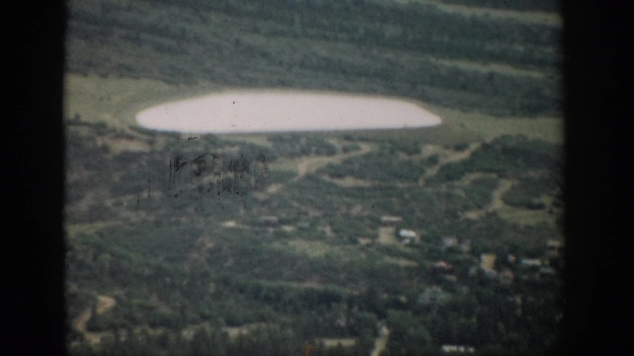 Soft aerial view of a pale lake beyond wooded hills and scattered small buildings.