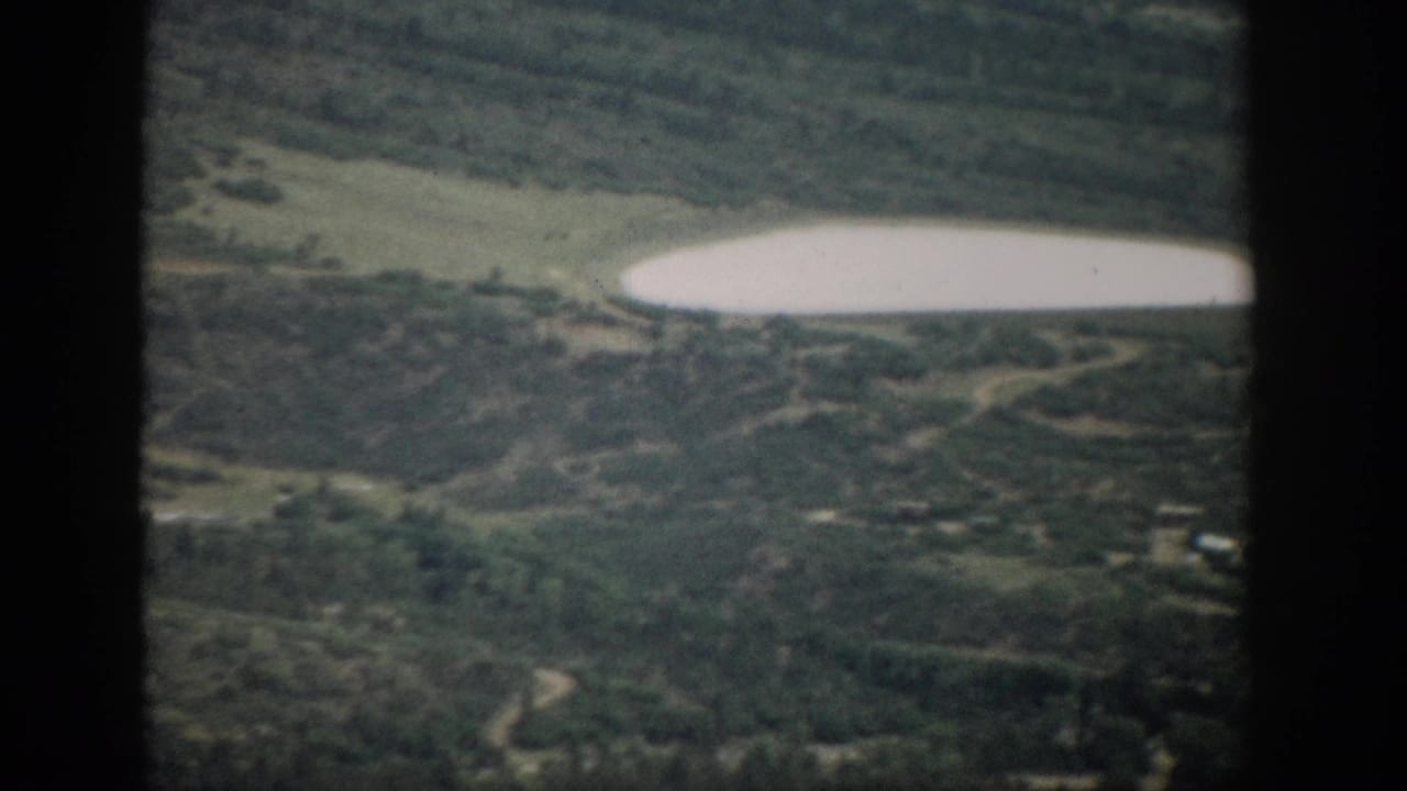 1959 Colorado home-movie frame with a bright lake, wooded slopes, faint roads, and scattered rural structures seen from above.