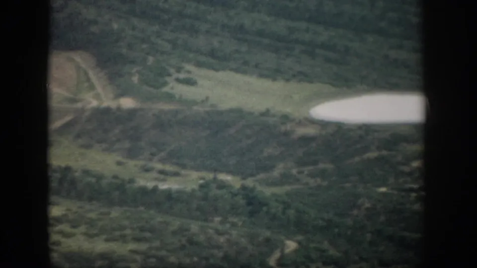 1959 Colorado 8mm frame showing part of a lake, surrounding forest, open ground, and winding roads from an elevated viewpoint.