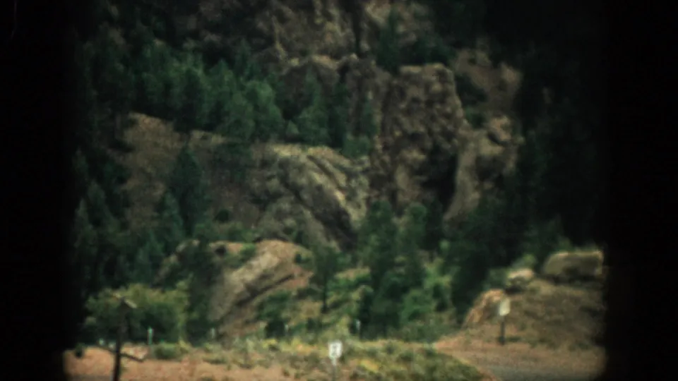 1959 Colorado 8mm frame showing evergreen trees, layered rocks, and dry hillside vegetation in bright sun.