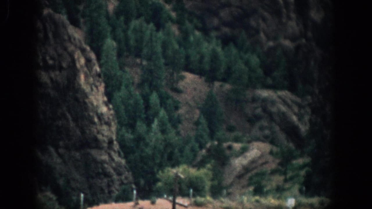 1959 Colorado 8mm frame showing rocky slopes, dense evergreens, and a dark window or film-frame edge at left.