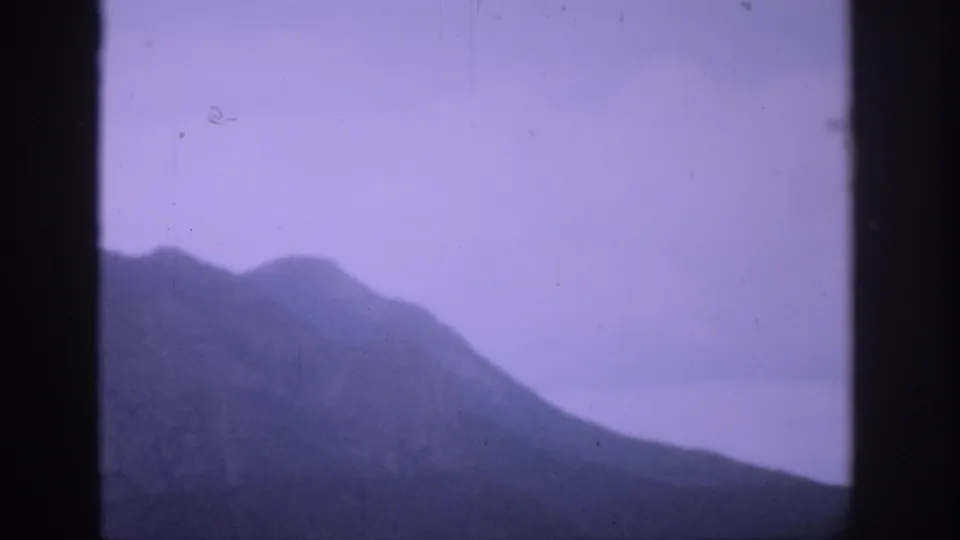 1959 Colorado 8mm mountain view with a shadowed ridge, pale purple sky, haze, and dark vertical frame borders.
