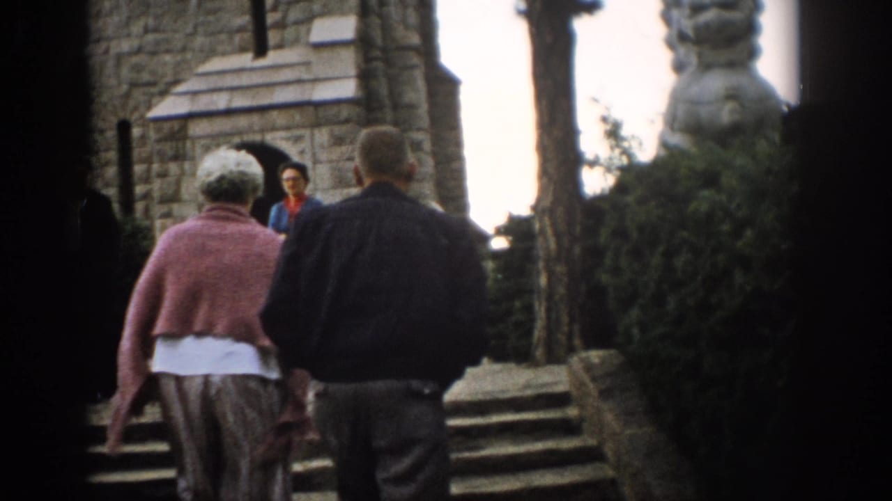 A woman in a pink cardigan and a man in a dark jacket ascend stone steps toward a Colorado church in 1959 footage.