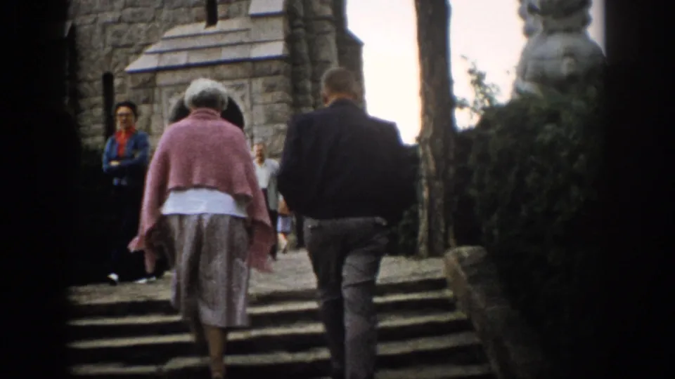The couple is viewed from behind on church steps, with other adults near the stone entrance in 1959 Colorado home-movie footage.