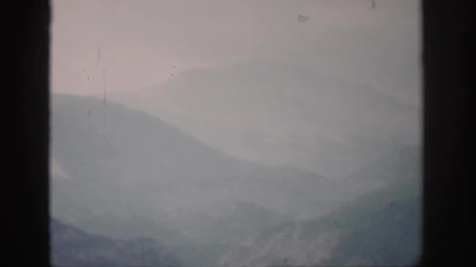 1959 Colorado home-movie frame of layered mountain slopes softened by fog, with muted color, dust, and slight scratches visible.
