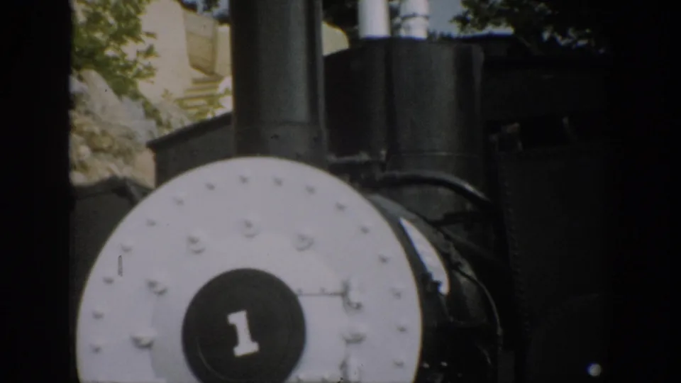 1959 Colorado 8mm frame showing the locomotive nose cropped low in frame, with rocky terrain and trees visible behind the engine.
