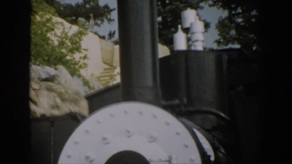 1959 Colorado 8mm frame with the steam locomotive front partly cut off, showing black metal cylinders, rocks, and trees in sunlight.