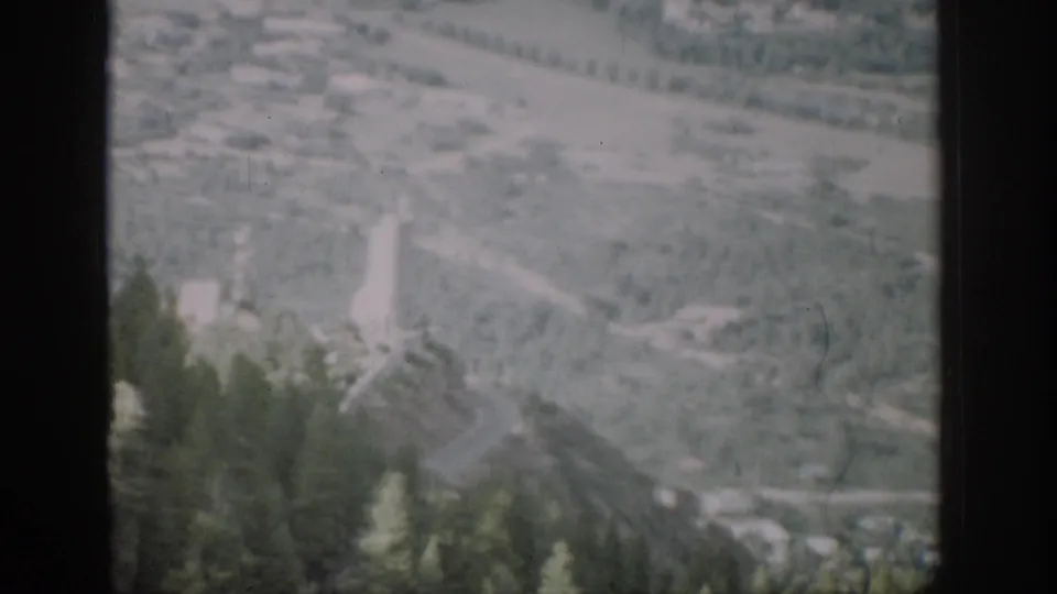 1959 Colorado 8mm frame looking from the train toward a mountain valley with trees, a winding road, and distant structures.