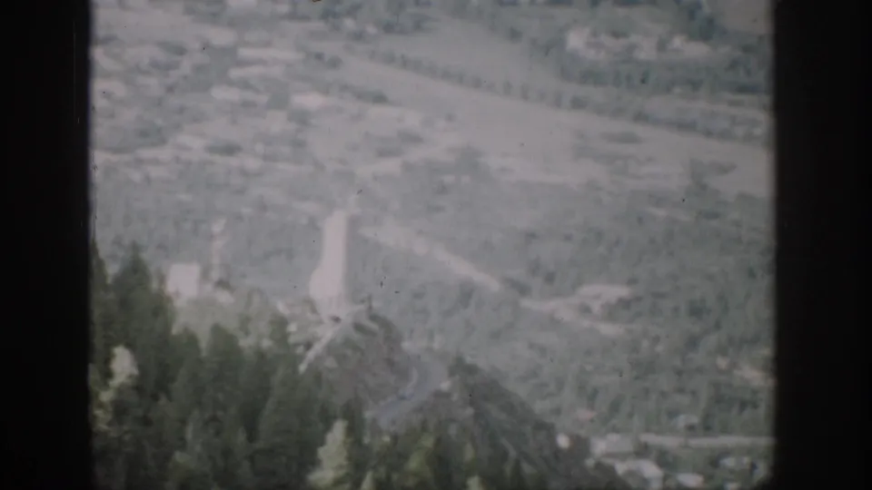 1959 Colorado home-movie frame with evergreen treetops below a hazy valley, winding road, and small pale structures.
