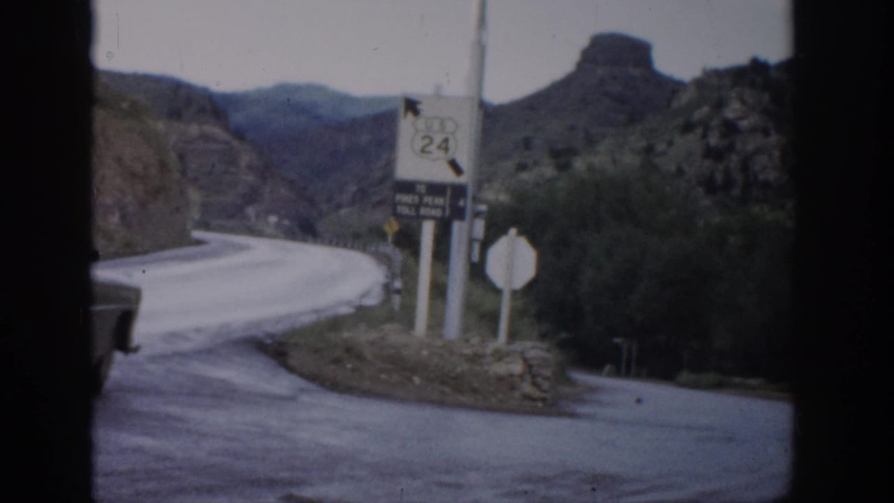 1959 Colorado 8mm frame showing a curving mountain road, Route 24 sign, toll-road marker, scrub vegetation, and rocky hills.