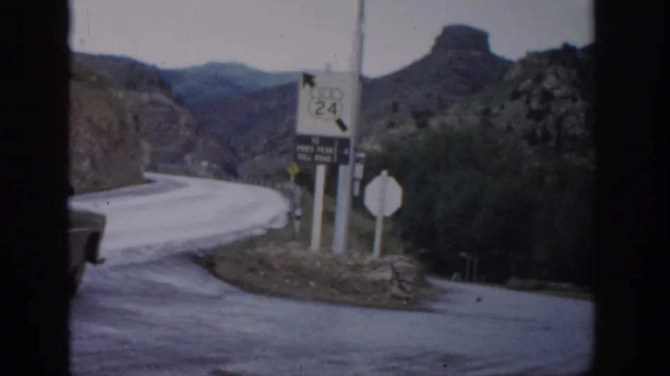1959 Colorado 8mm frame showing a curving mountain road, Route 24 sign, toll-road marker, scrub vegetation, and rocky hills.