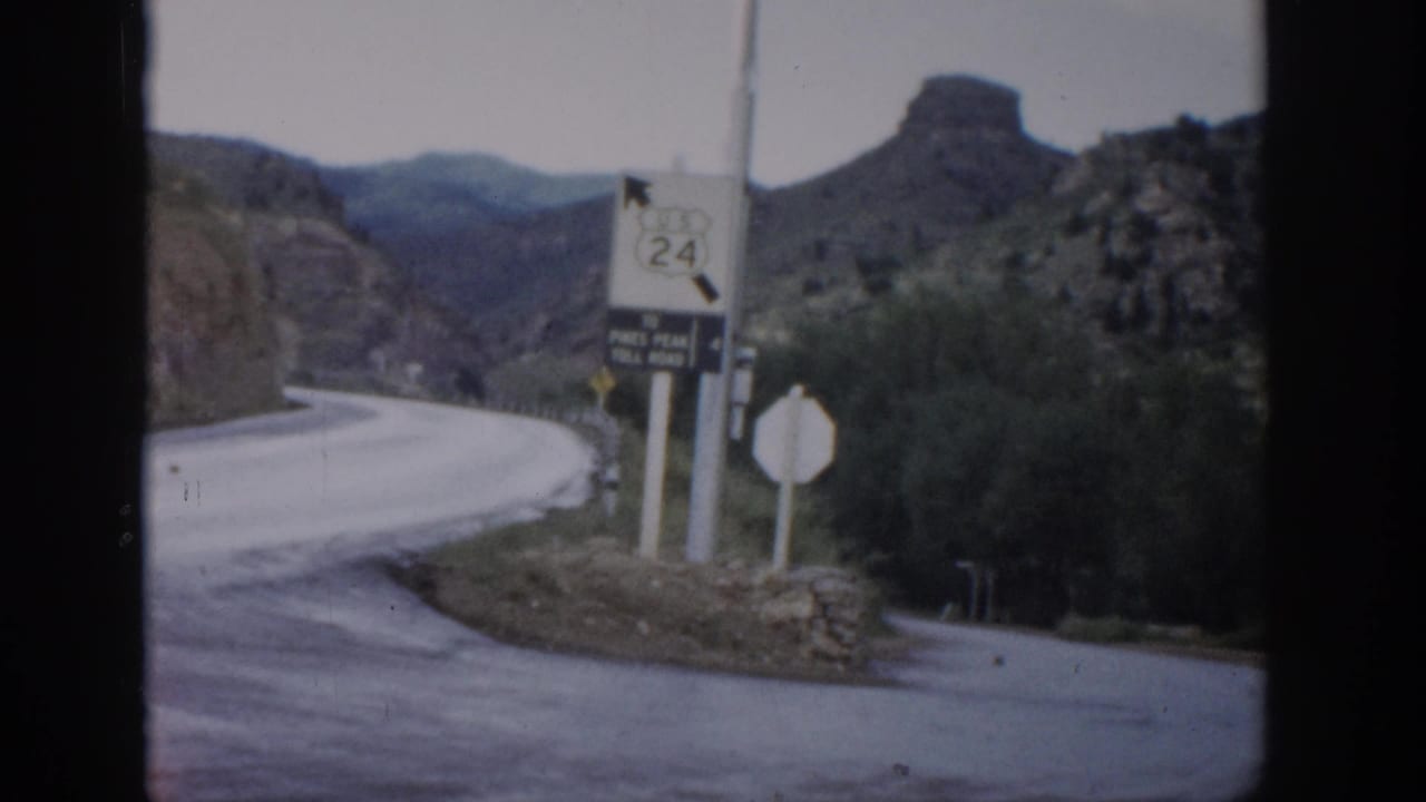 1959 Colorado frame with Route 24 and toll-road signage beside a winding road, low brush, rock formations, and distant mountains.
