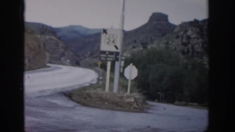 1959 Colorado frame with Route 24 and toll-road signage beside a winding road, low brush, rock formations, and distant mountains.