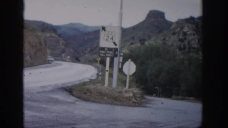 1959 Colorado 8mm still showing a highway curve, Route 24 sign, toll-road marker, brush, guardrail, and mesa-like rock forms.