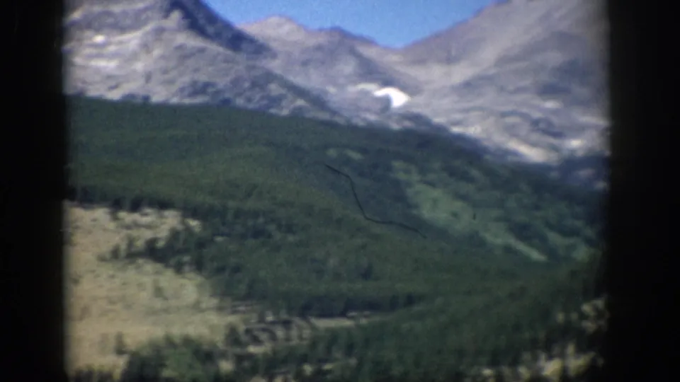 Soft 8mm Colorado landscape frame showing a green valley, conifer forest, distant ridges, and lingering snow.