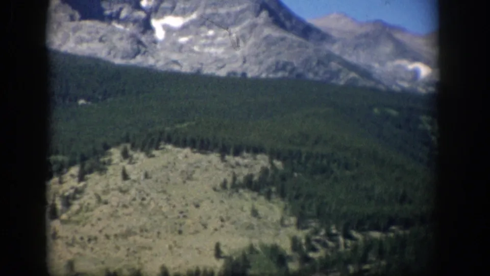 Final 1959 Colorado frame with rocky foreground, dense conifers, gray mountain ridges, and patches of snow near the top.