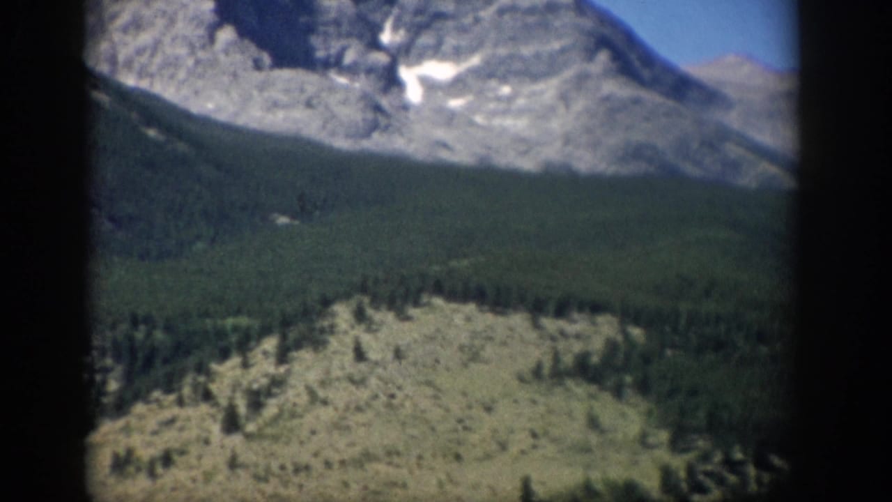 1959 Colorado 8mm frame showing gray mountain peaks, patches of snow, dark evergreen forest, and sunlit scrubland below.