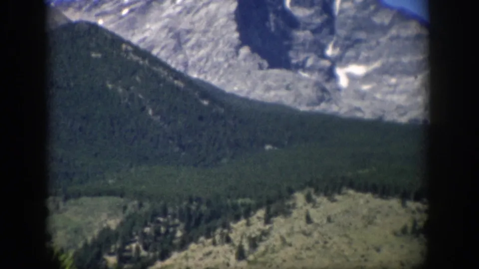 1959 Colorado home-movie frame with a darker forested ridge, pale rock above, snow patches, and soft handheld blur.