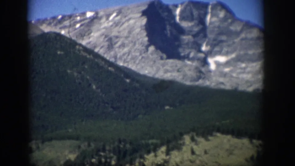 1959 Colorado 8mm frame focused on rocky snow-patched peaks above a dark green forested slope and lower meadow.