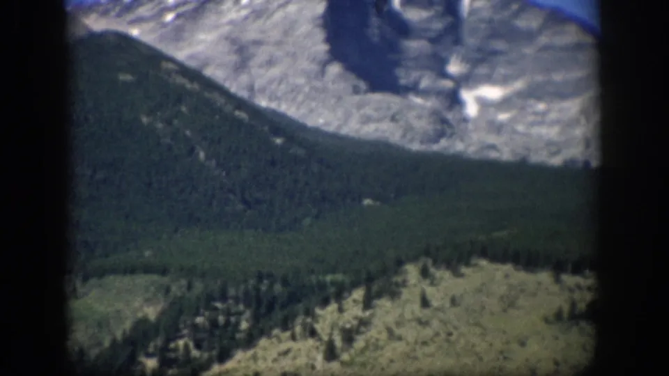 1959 Colorado 8mm frame showing a pale mountain peak, dark forested ridge, and rocky hillside within projector borders.
