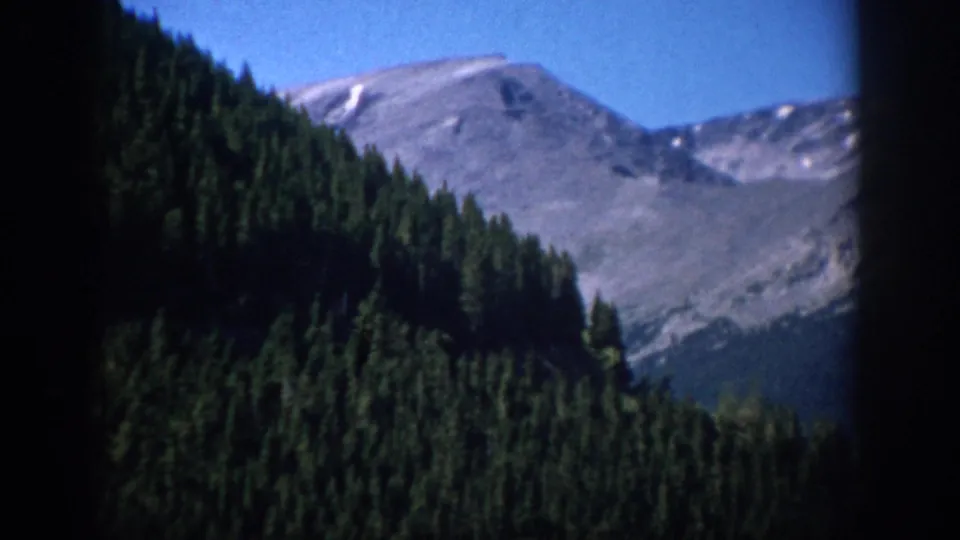 1959 Colorado landscape frame showing dense evergreen forest rising toward a gray mountain ridge under blue sky.