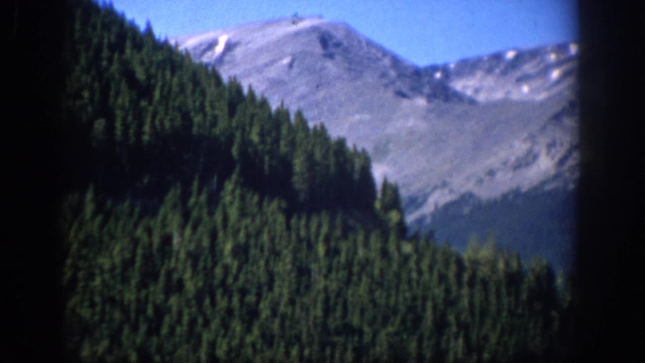 1959 Colorado 8mm home-movie frame showing evergreen-covered slopes beneath rocky mountain peaks and a clear blue sky.