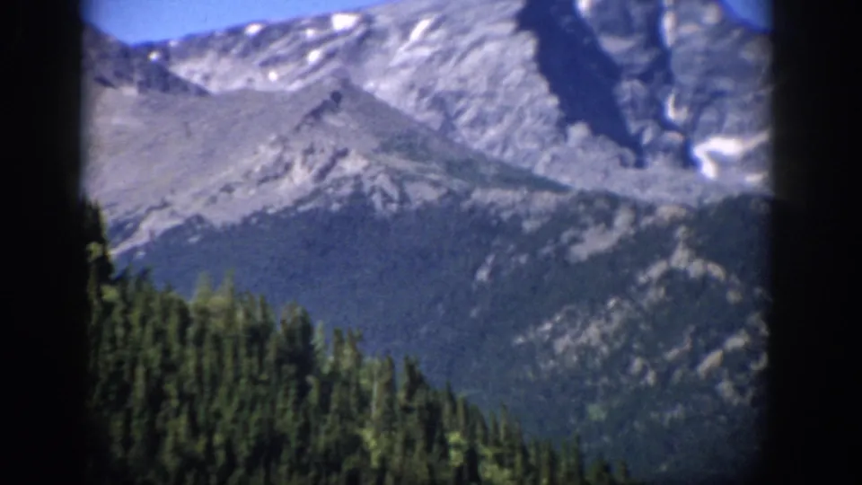 1959 Colorado home-movie still of a rocky mountain face with remnant snow above dark evergreen forest.