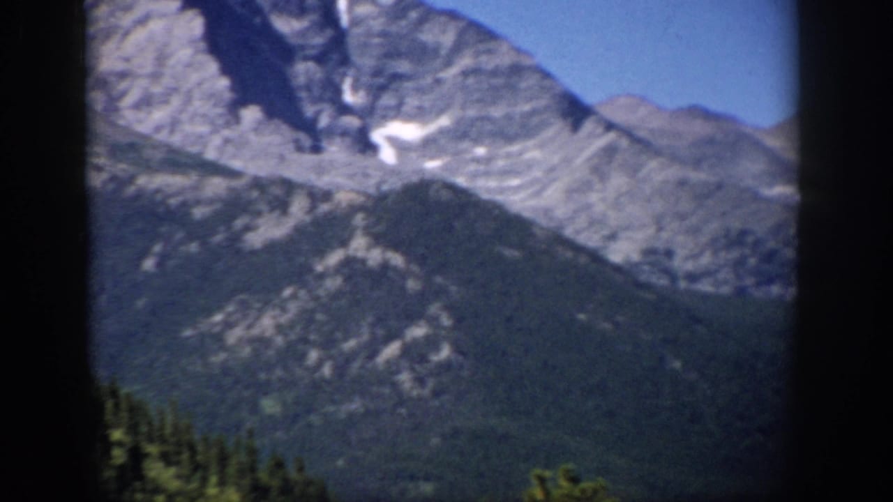 1959 Colorado 8mm frame showing a rugged mountain peak, lower forested slopes, small snow patches, and dark film borders.