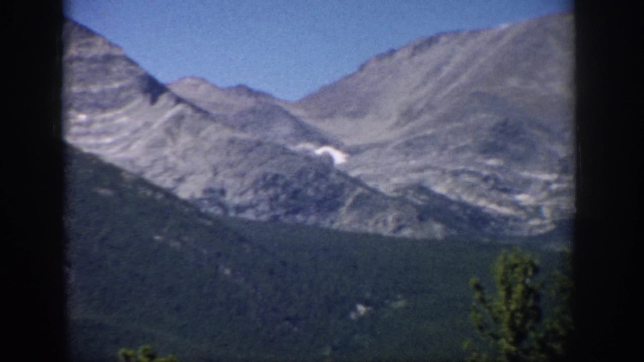 Broad gray mountain ridge above dark green forest with a small snow patch and blue sky.