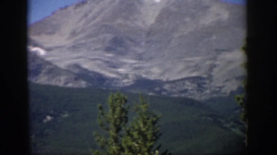 1959 Colorado 8mm frame with a rocky mountain slope, dark forested base, foreground evergreen treetops, and visible film softness.
