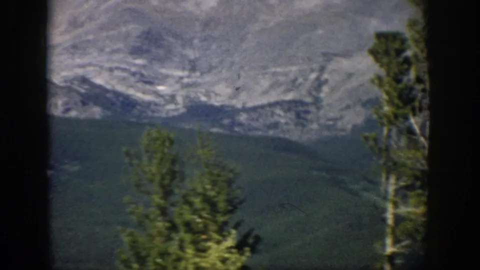 1959 Colorado 8mm view of a rocky mountain face above forested lower slopes, with evergreen trees at the right foreground.