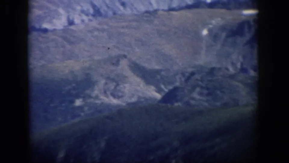 1959 Colorado 8mm frame of rugged mountain slopes, pale snow patches, and deep blue-green shadows within dark borders.