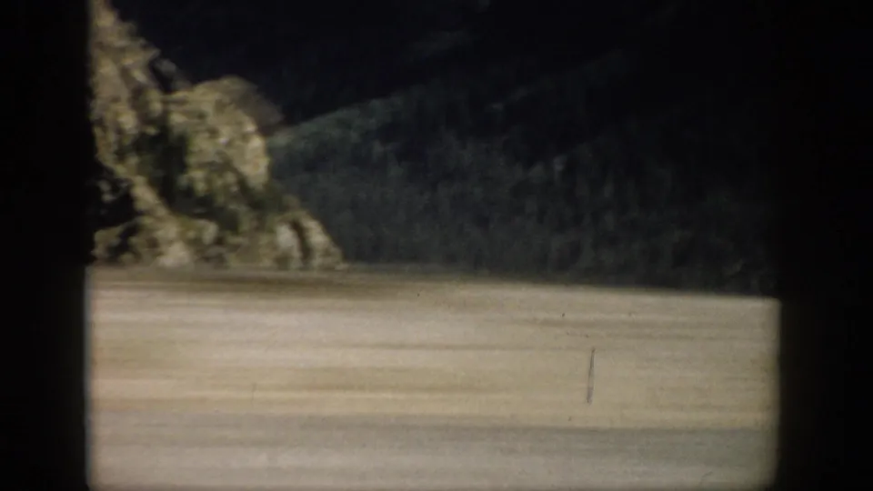 1959 Colorado 8mm view of a rocky slope beside a pale open foreground, with forested mountain terrain in the distance.