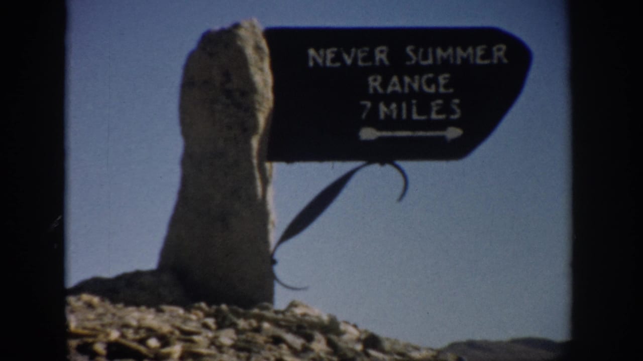 Black Never Summer Range 7 Miles sign mounted beside a rough stone post under blue sky.