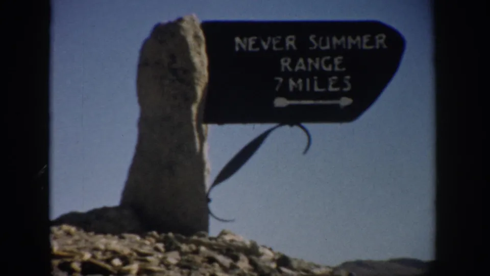 1959 Colorado 8mm frame showing a black directional sign reading Never Summer Range 7 Miles beside a weathered stone post.