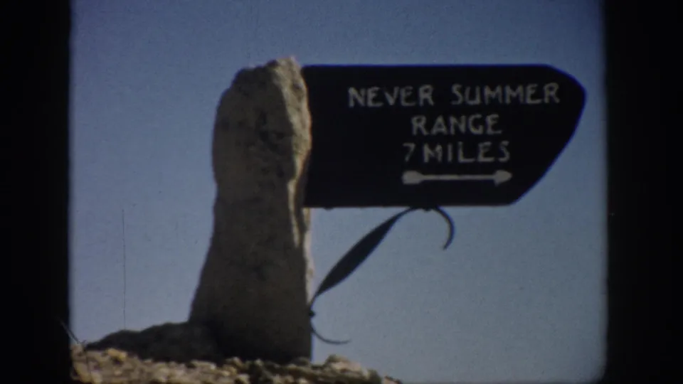Tight 1959 Colorado home-movie view of the Never Summer Range 7 Miles sign with a rough stone post and open sky.