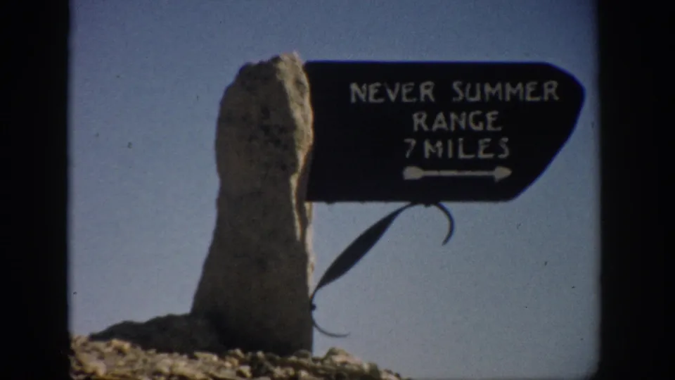 Color 8mm frame from Colorado showing the Never Summer Range sign beside a tall stone marker, with rocky ground below.