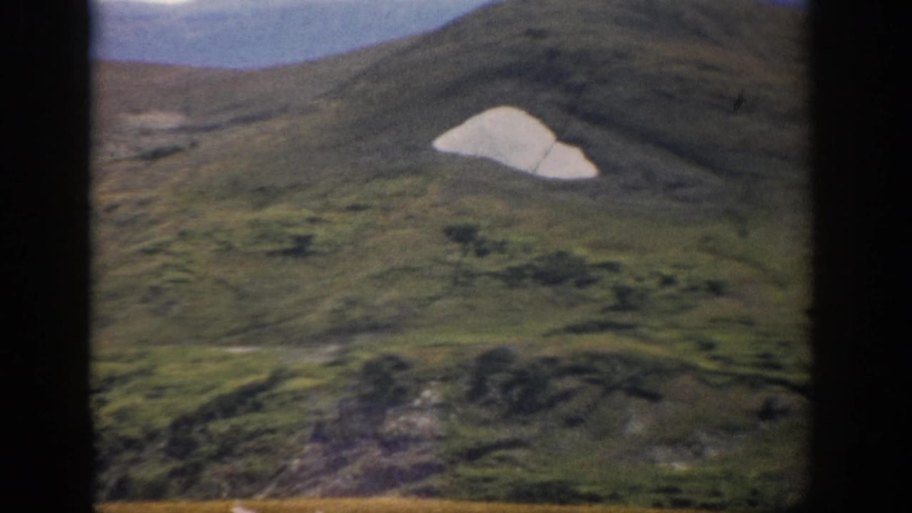 1959 Colorado 8mm frame showing a green mountainside with a pale snow or rock patch, soft sky, and visible film-frame edges.
