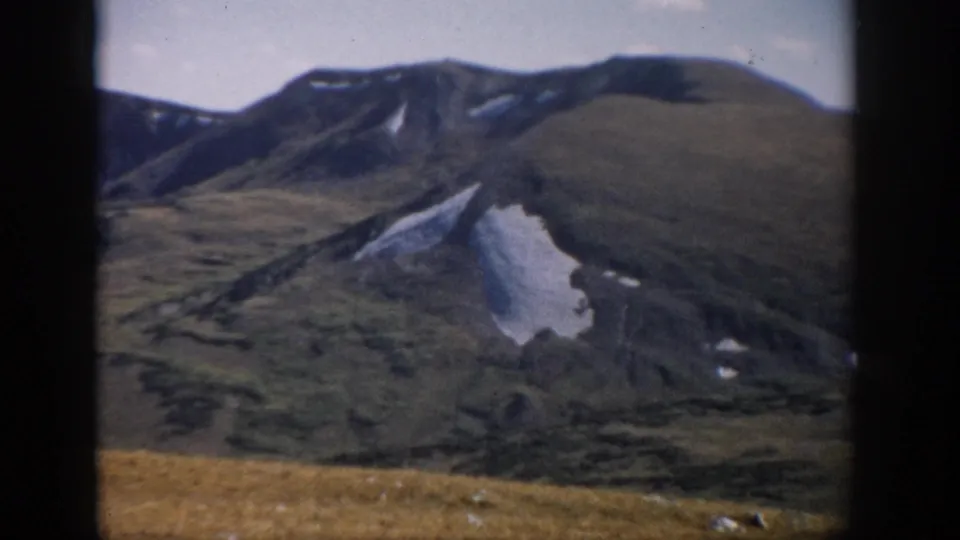 1959 Colorado mountain landscape with golden grass foreground, green slopes, dark ridges, and patches of snow below a pale sky.