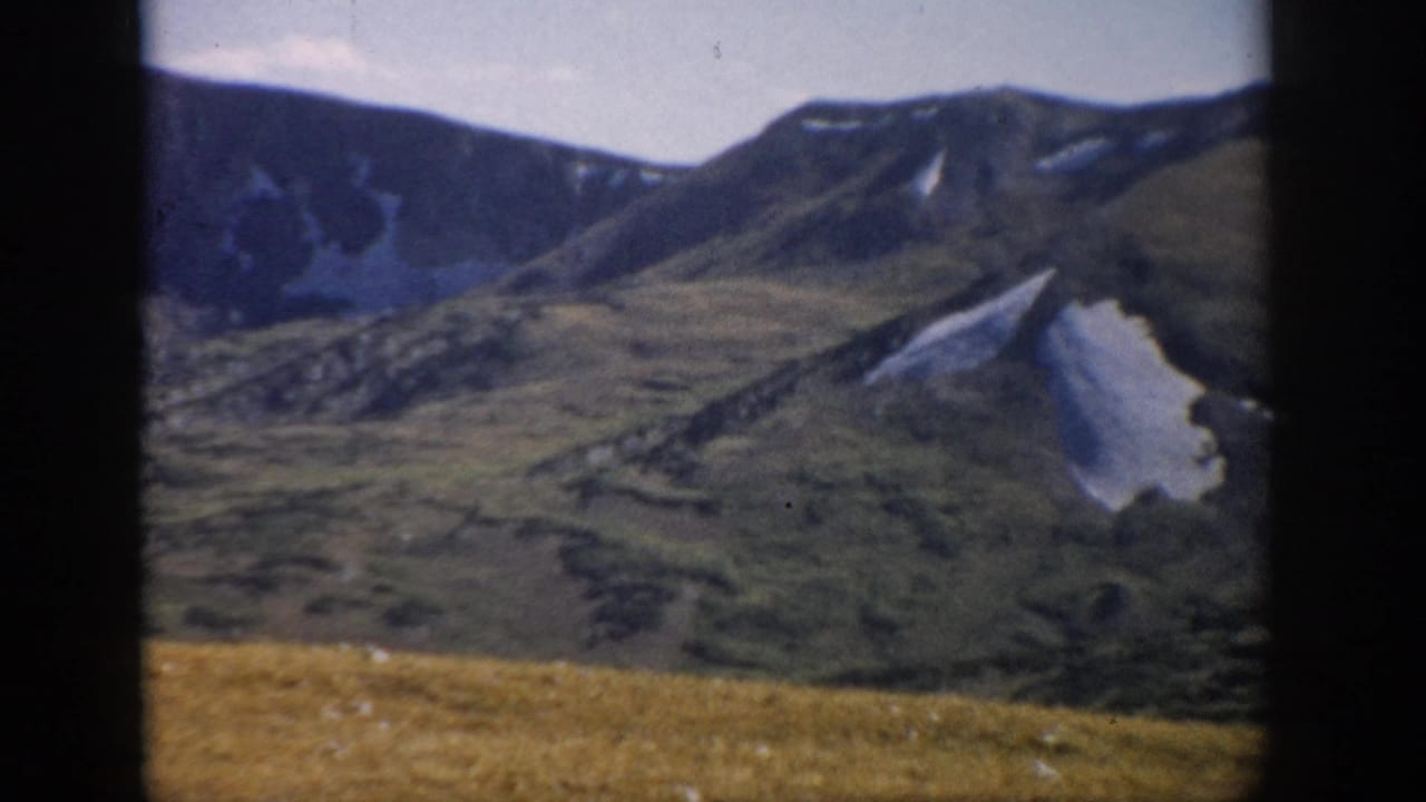1959 Colorado 8mm view of rugged mountain slopes and snow patches beyond a high alpine meadow, seen through dark frame edges.