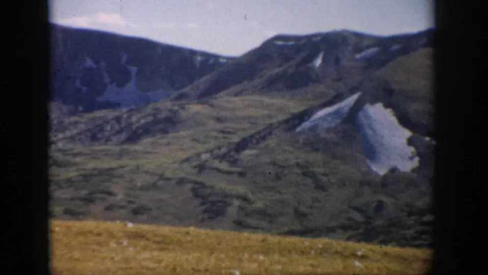 1959 Colorado 8mm view of rugged mountain slopes and snow patches beyond a high alpine meadow, seen through dark frame edges.