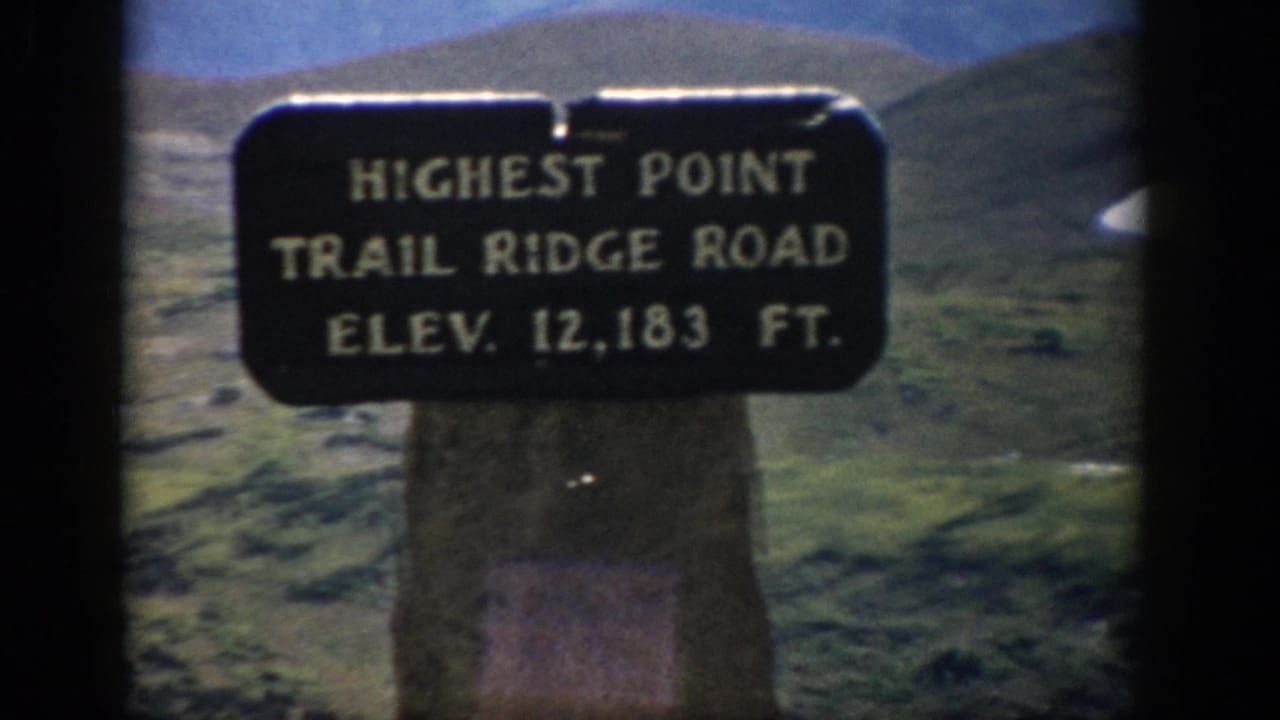 1959 Colorado 8mm frame showing the Trail Ridge Road highest point elevation sign mounted on a stone post in open alpine terrain.