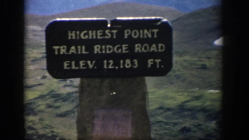 1959 Colorado 8mm frame showing the Trail Ridge Road highest point elevation sign mounted on a stone post in open alpine terrain.
