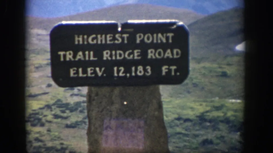 Visitor-shot 1959 Colorado frame with the 12,183-foot Trail Ridge Road sign centered before rugged high-country slopes.